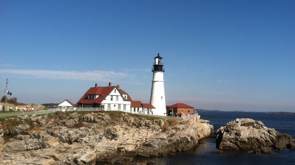 A white lighthouse stands on a rocky cliff beside a house with a red roof in Portland Maine.