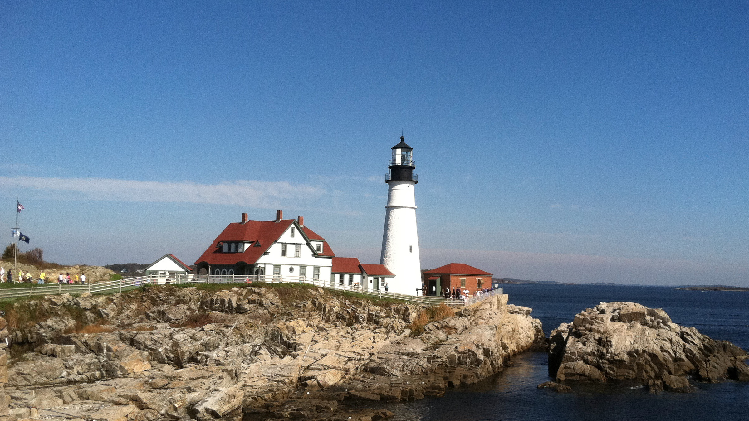 A white lighthouse stands on a rocky cliff beside a house with a red roof in Portland Maine.