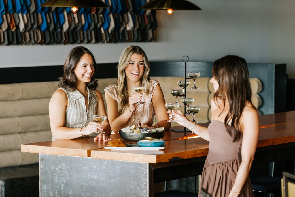 Three women laughing and drinking cocktails at a wooden bar in a restaurant in Portland Maine.
