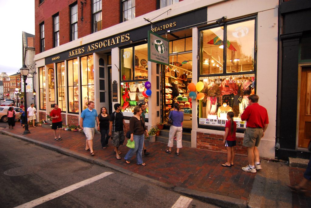 People walking on a brick sidewalk past a storefront with large display windows in Portland, Maine. Photo Credit: Chris Lawrence