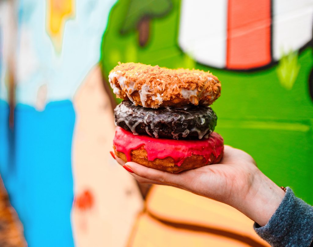 A hand holding a stack of three colorful glazed donuts against a mural background in Portland Maine.