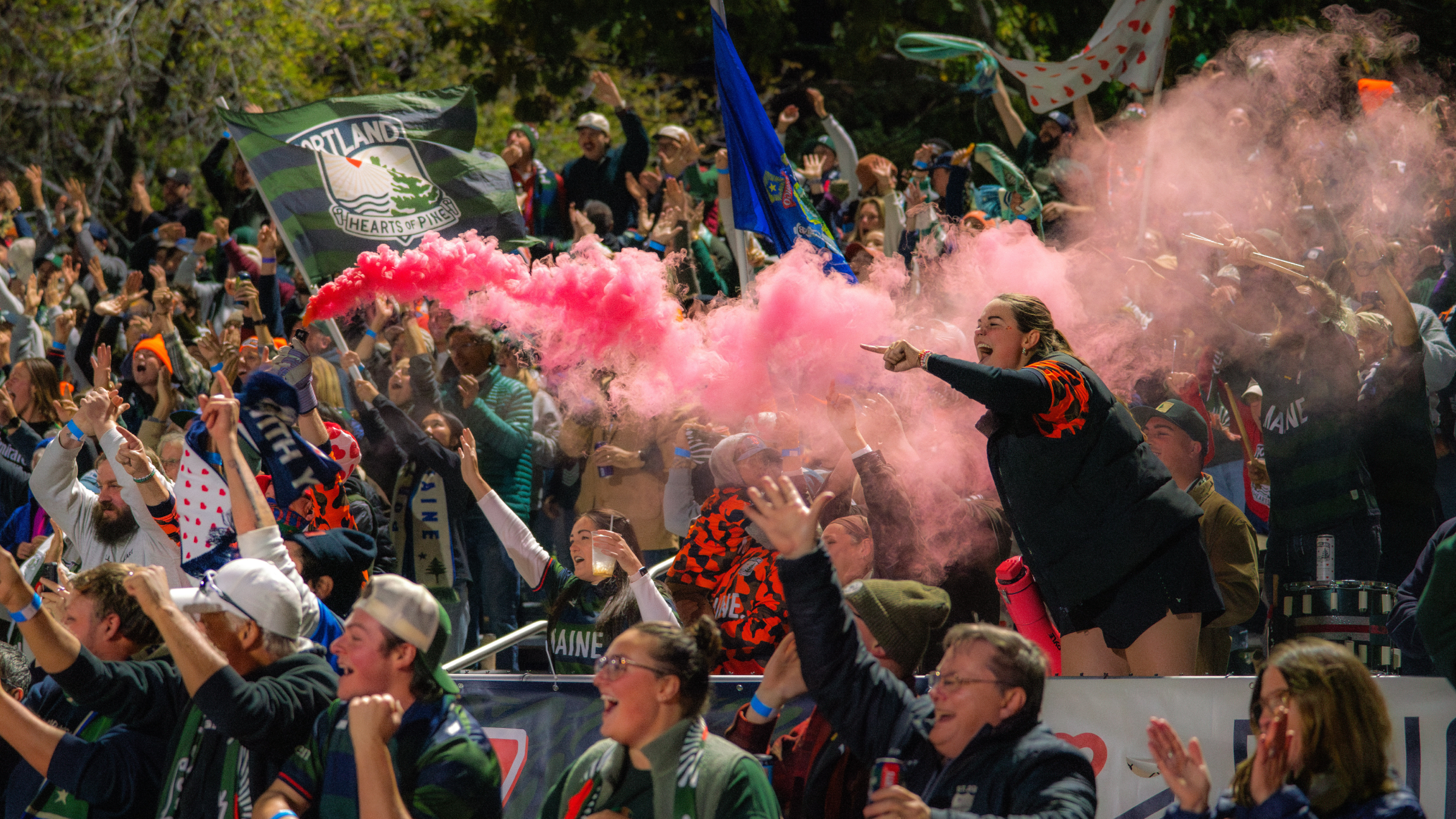 A crowd of soccer fans cheering with pink smoke and flags in a stadium in Portland Maine.