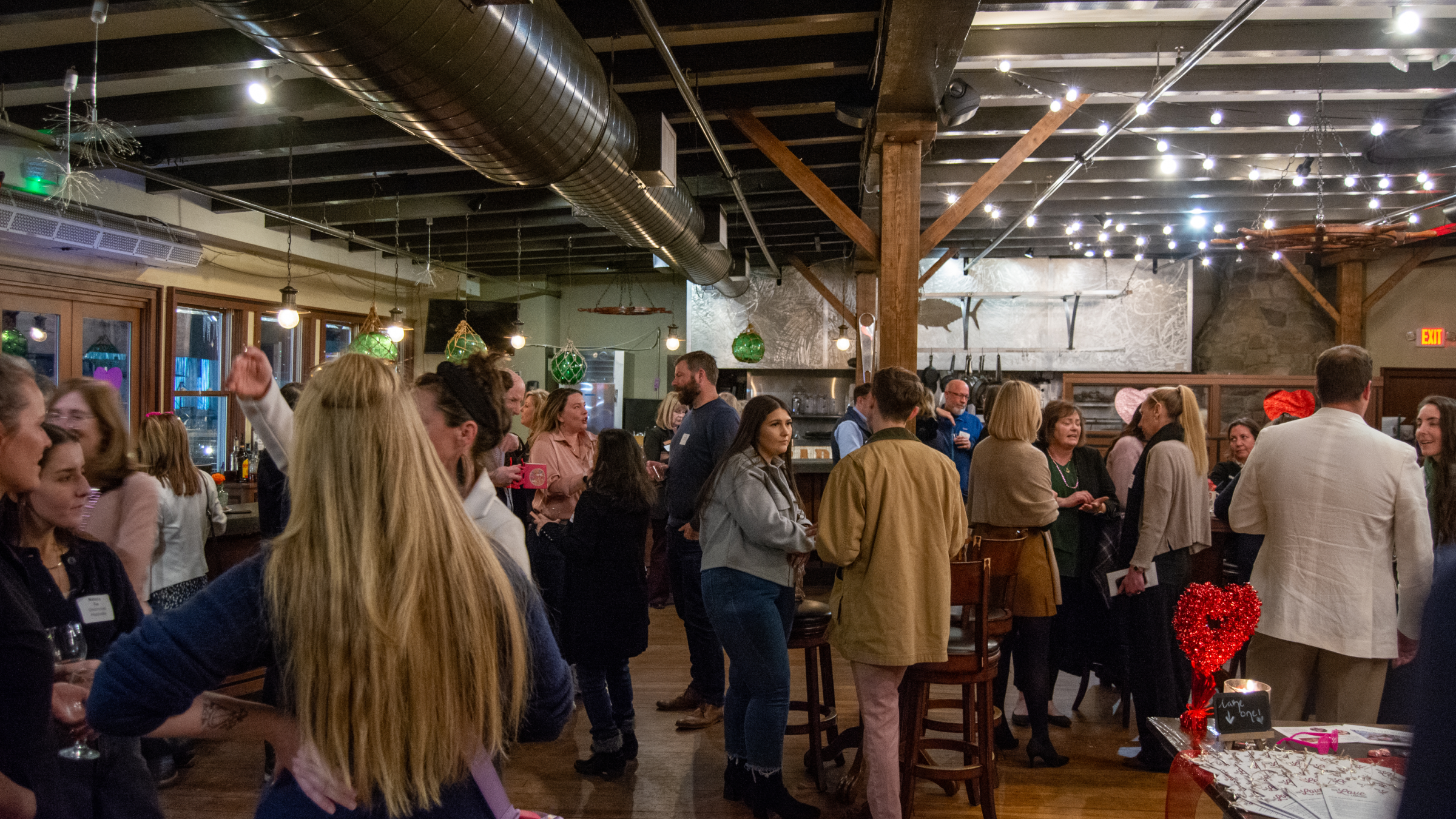 A group of people socializing at an indoor event with string lights and wooden beams in Portland Maine.