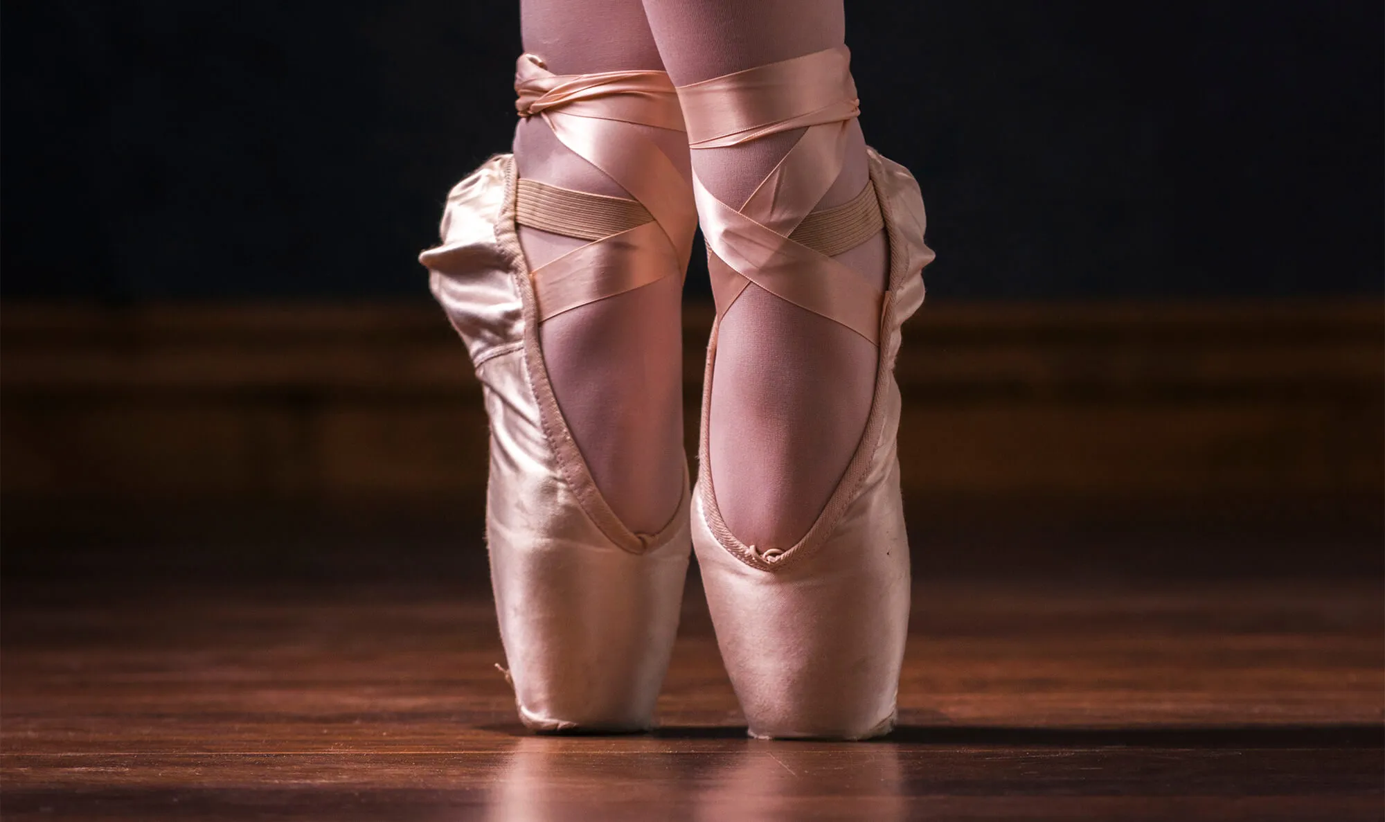 A close-up of a ballerina's feet in pink pointe shoes on a wooden floor in Portland Maine.