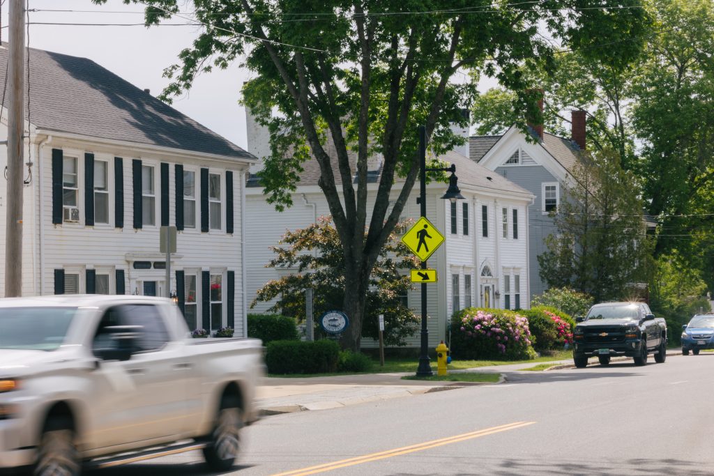 A white truck drives past historic houses on a sunny street with a pedestrian sign in Portland Maine.