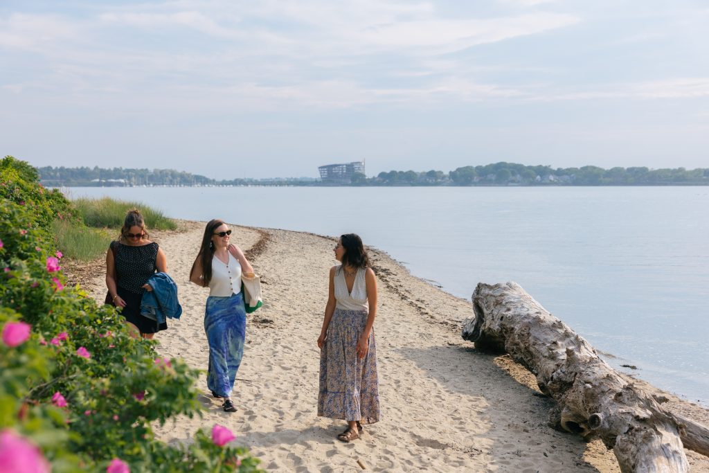Three women walking along a sandy beach near pink flowers and a large log in Portland Maine.