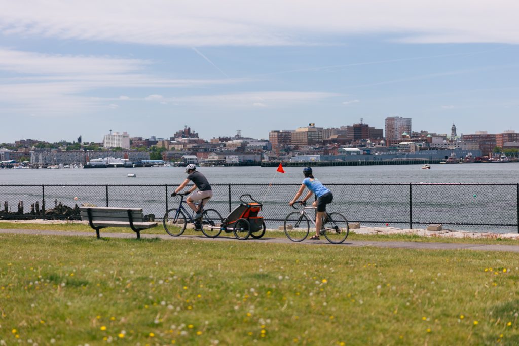 Two people riding bicycles along a waterfront path with a city skyline in the background in Portland Maine.