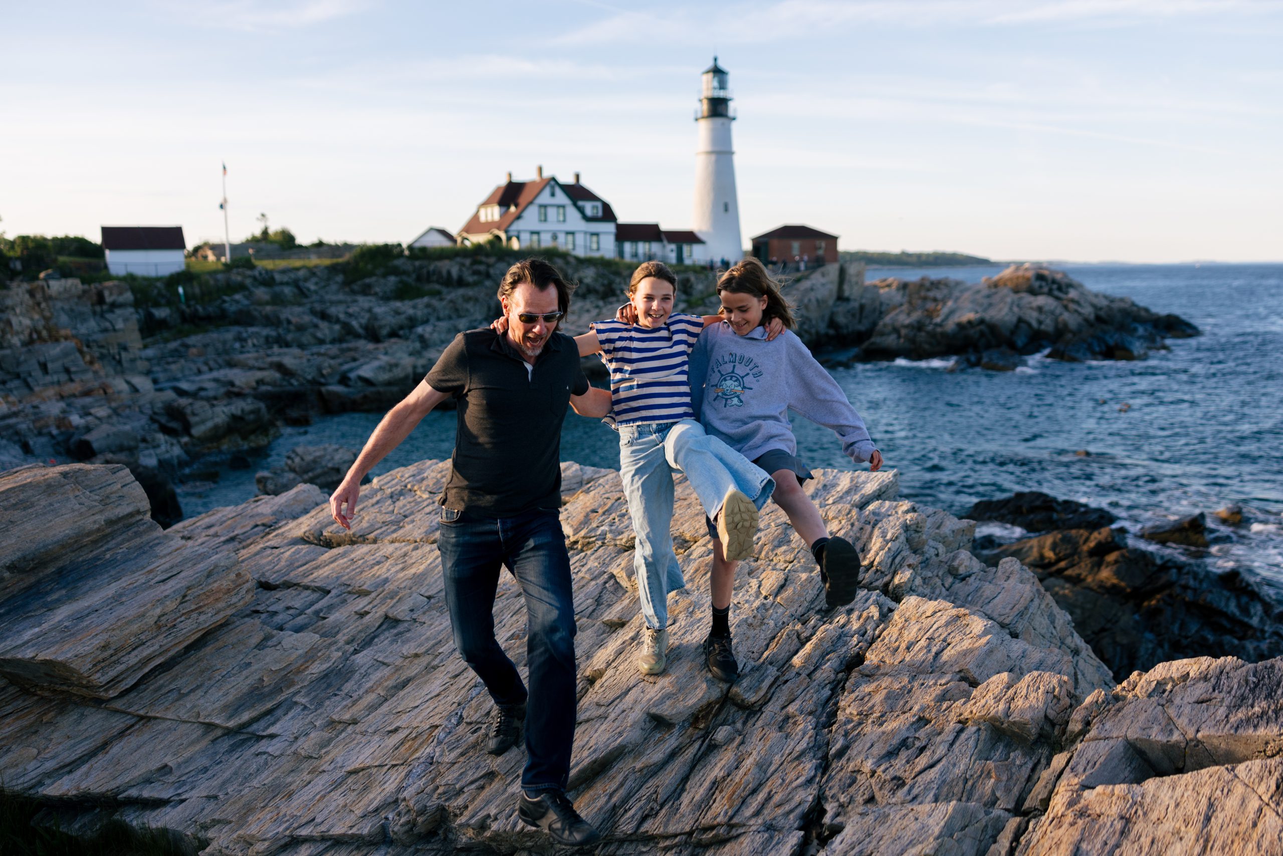 A father and two children walking playfully on rocky coastal terrain near a lighthouse in Portland Maine.