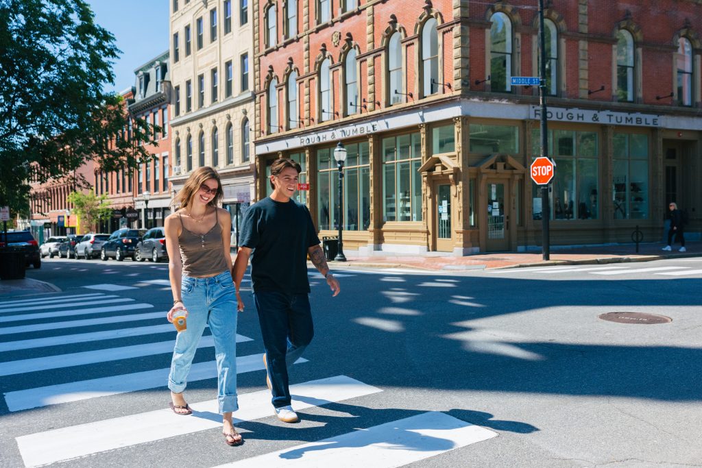 A young couple walks hand-in-hand across a city crosswalk on a sunny day in Portland Maine.