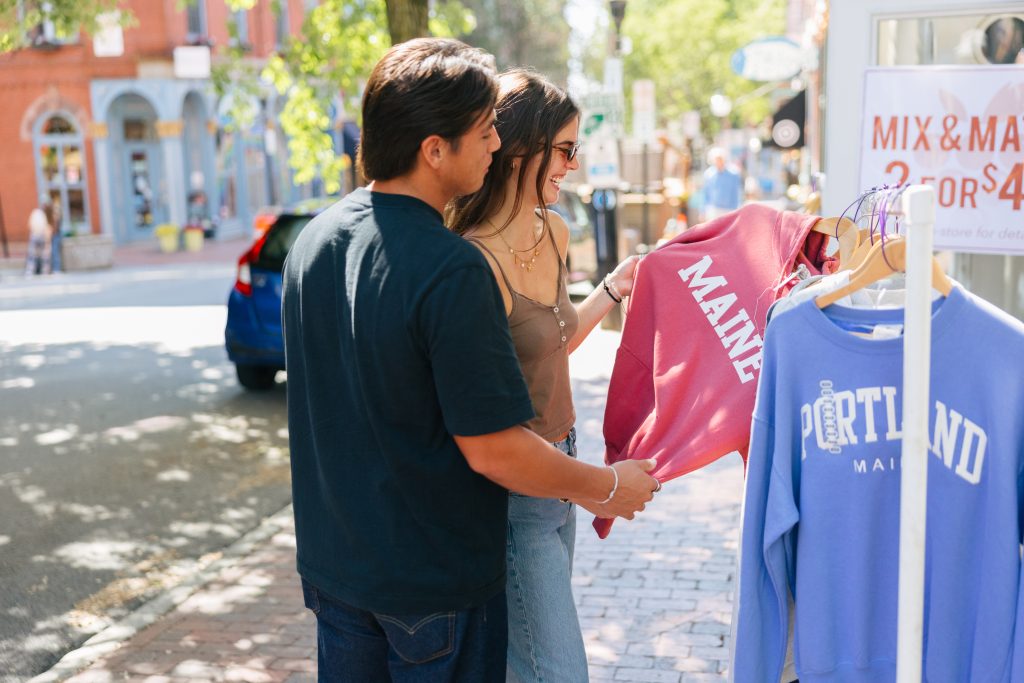A young couple shops for sweatshirts at an outdoor clothing rack on a sunny day in Portland Maine.